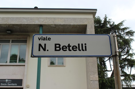 Targa stradale che indica viale N. Betelli, con un edificio sullo sfondo e alberi ai lati.