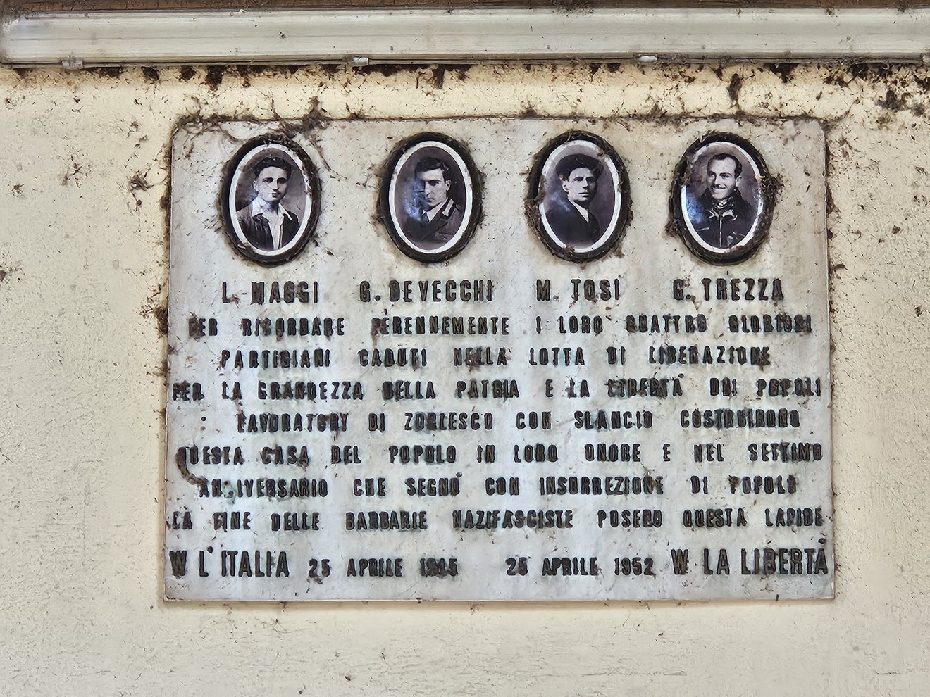 Lapide in marmo su parete esterna, con quattro ritratti fotografici ovali e un'iscrizione.