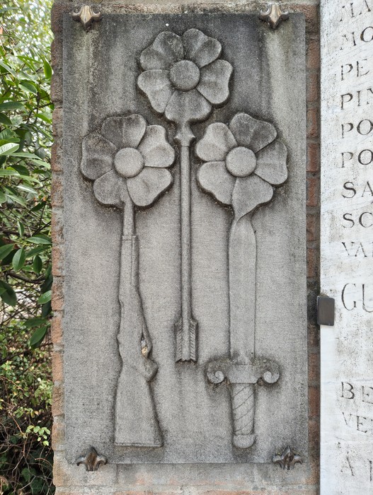 Lastra in pietra con tre fiori e tre armi scolpite a rilievo, fucile, spada e pugnale, accanto a un'iscrizione.