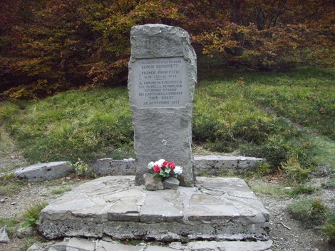 Cippo commemorativo dedicato a Giovannetti e Prioreschi, immerso nella natura, con fiori ai suoi piedi.