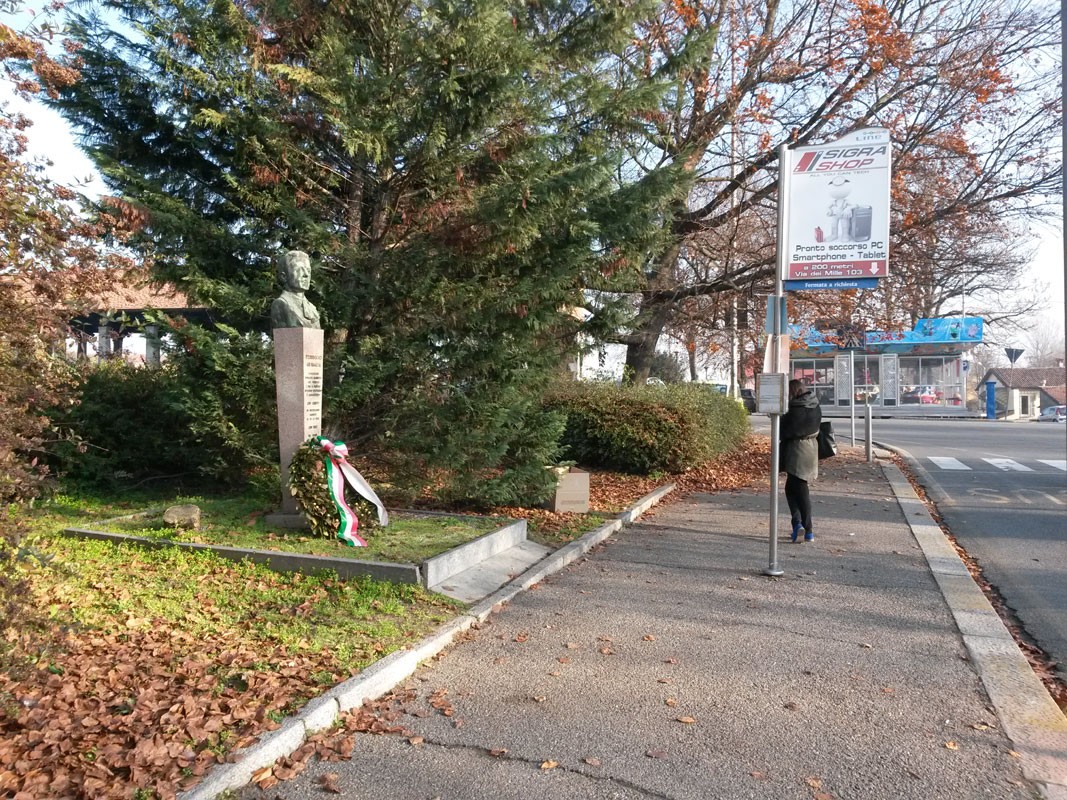 Monumento a Ghinaglia, un omaggio che si erge tra alberi e foglie secche, con una corona di fiori ai suoi piedi.