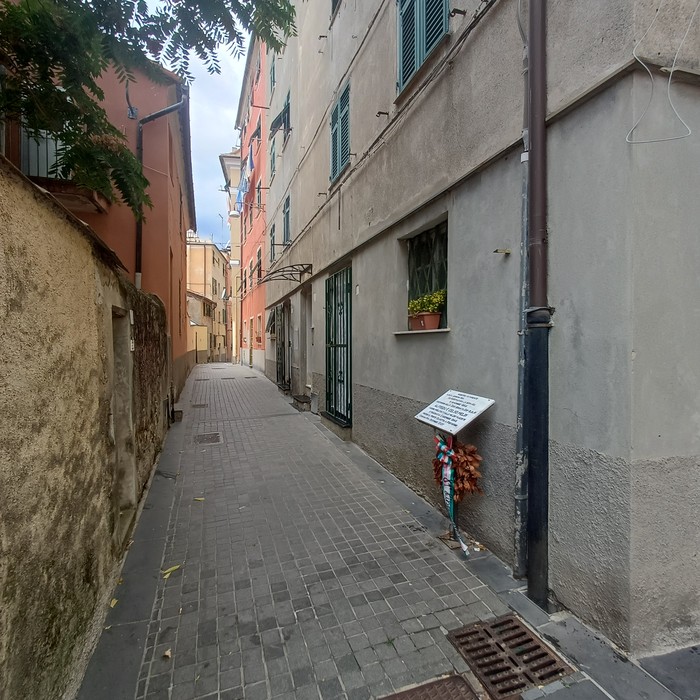 Lapide con iscrizione su parete di vicolo stretto, con fiori deposti alla base.