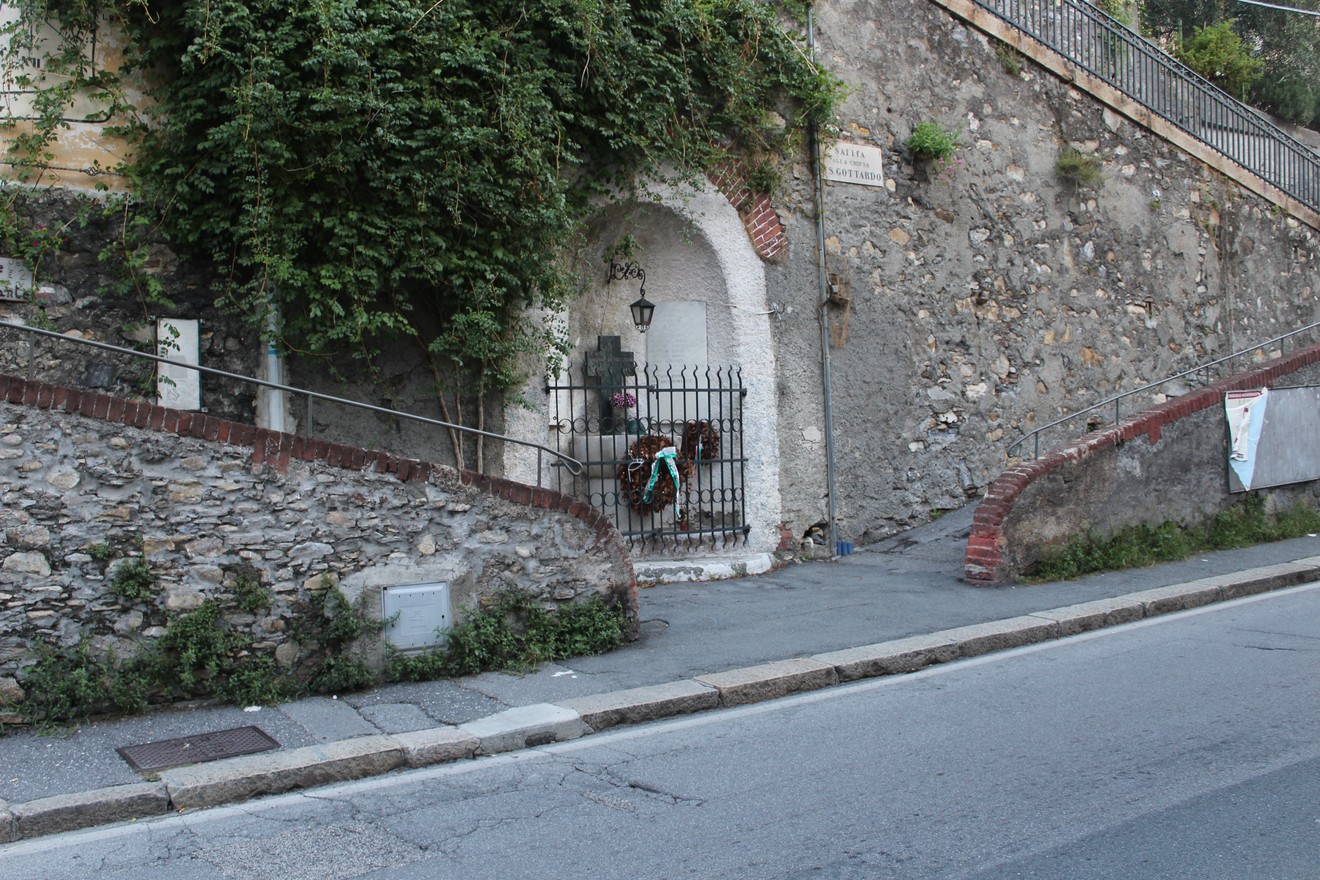 Edicola dei caduti di San Gottardo, affacciata su una strada, circondata da vegetazione e muretti in pietra.