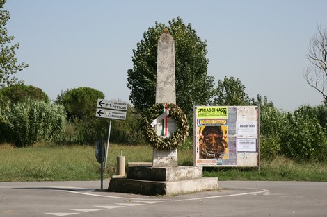 Monumento di Pettori: obelisco circondato da vegetazione, con una corona e manifesti nelle vicinanze.