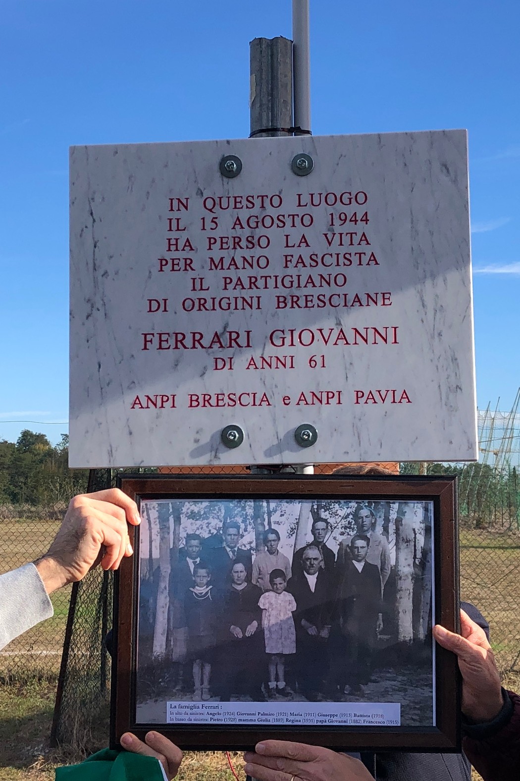 Lapide in marmo con iscrizione, fissata a una struttura metallica all’aperto; sotto è esposta una fotografia incorniciata.