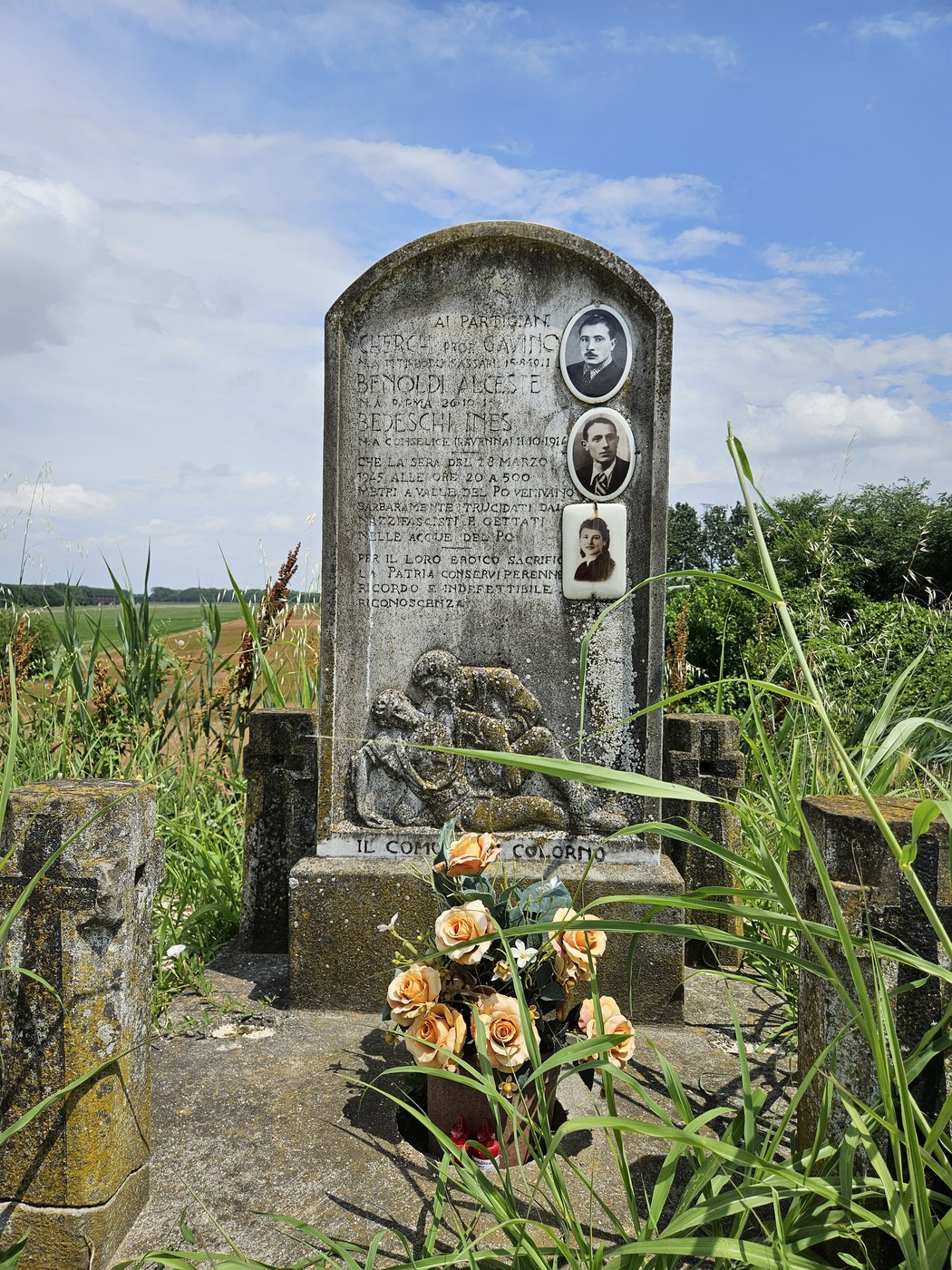 Cippo commemorativo in un'area rurale, circondato da vegetazione e fiori, dedicato a Cherchi, Benoldi e Bedeschi.