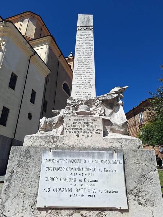 Monumento in pietra con obelisco, statue e lapide con iscrizione, in una piazza davanti a una chiesa.