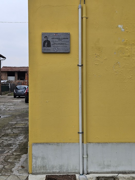 Targa commemorativa con ritratto e iscrizione, fissata sulla parete gialla di un edificio.
