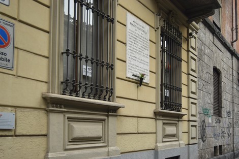 Lapide commemorativa affissa su una parete di un edificio, con finestre in ferro battuto e muratura storica.