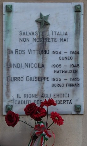 Lapide commemorativa in marmo con nomi e date di tre persone, decorata con rose rosse.