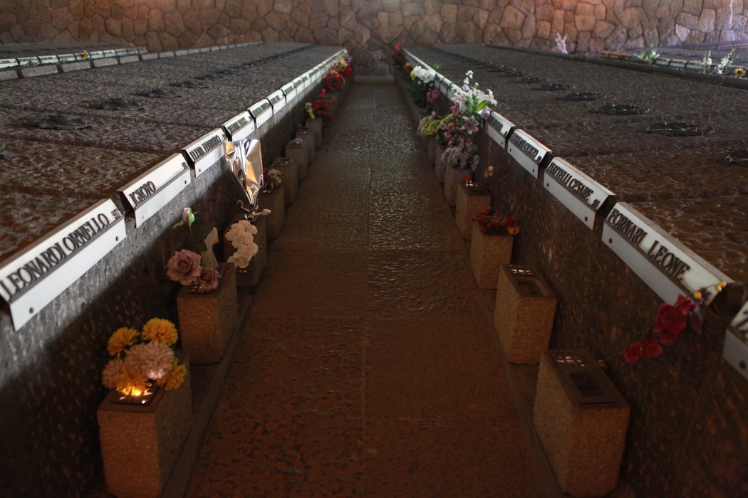 Interno del Mausoleo delle Fosse Ardeatine, con loculi e fiori commemorativi. Atmosfera solenne e rispettosa.