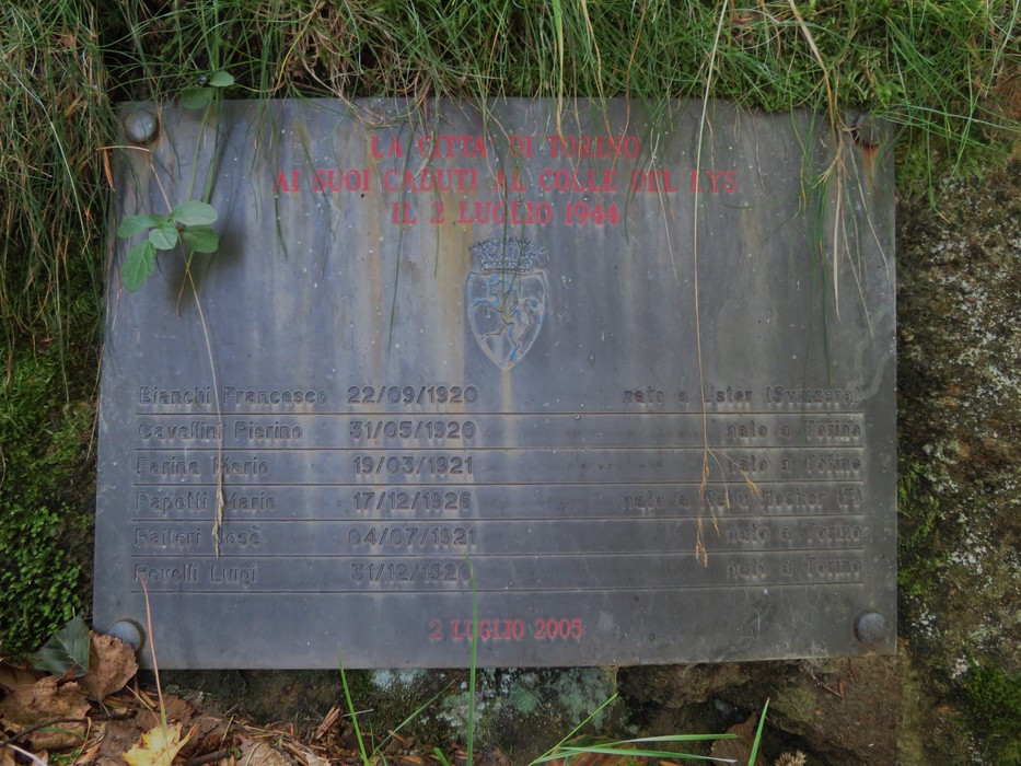Memoriale di una fossa comune con targa commemorativa e nomi delle persone scomparse. Ambiente naturale circostante.