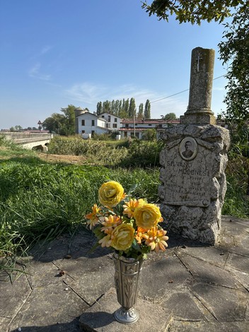 Cippo commemorativo a Modenesi, circondato da fiori e un paesaggio verdeggiante.