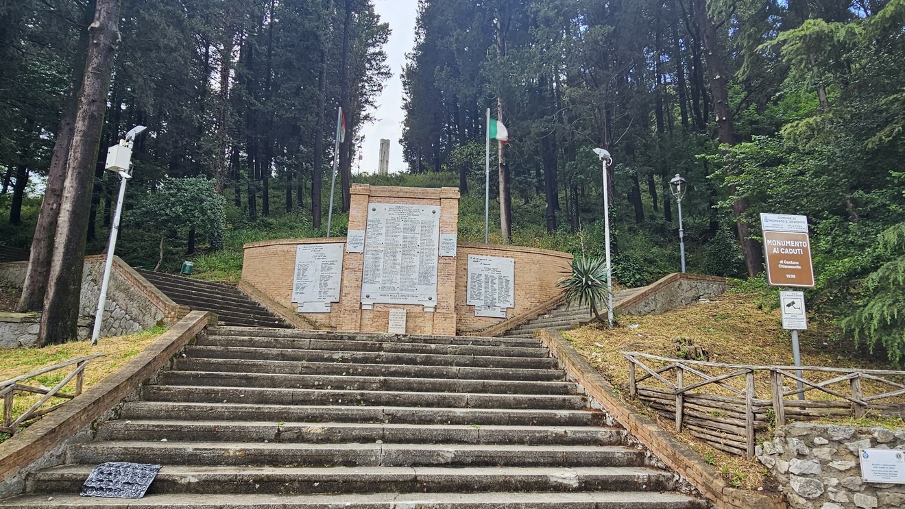 Monumento ai caduti circondato da alberi, con scalinate e pannelli commemorativi visibili.