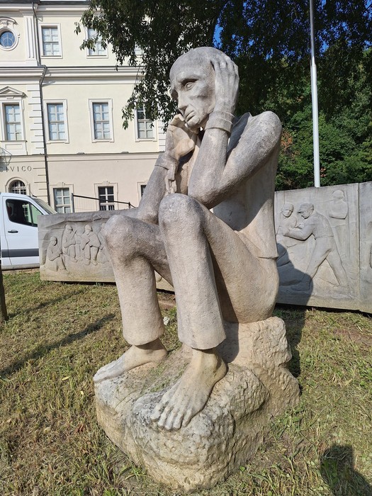 Statua in pietra di un uomo seduto su una roccia, a piedi nudi, con il viso appoggiato alle mani.