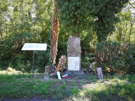 Cippo in pietra con lapide e iscrizione, tra alberi, con fiori e nastro tricolore ai piedi.