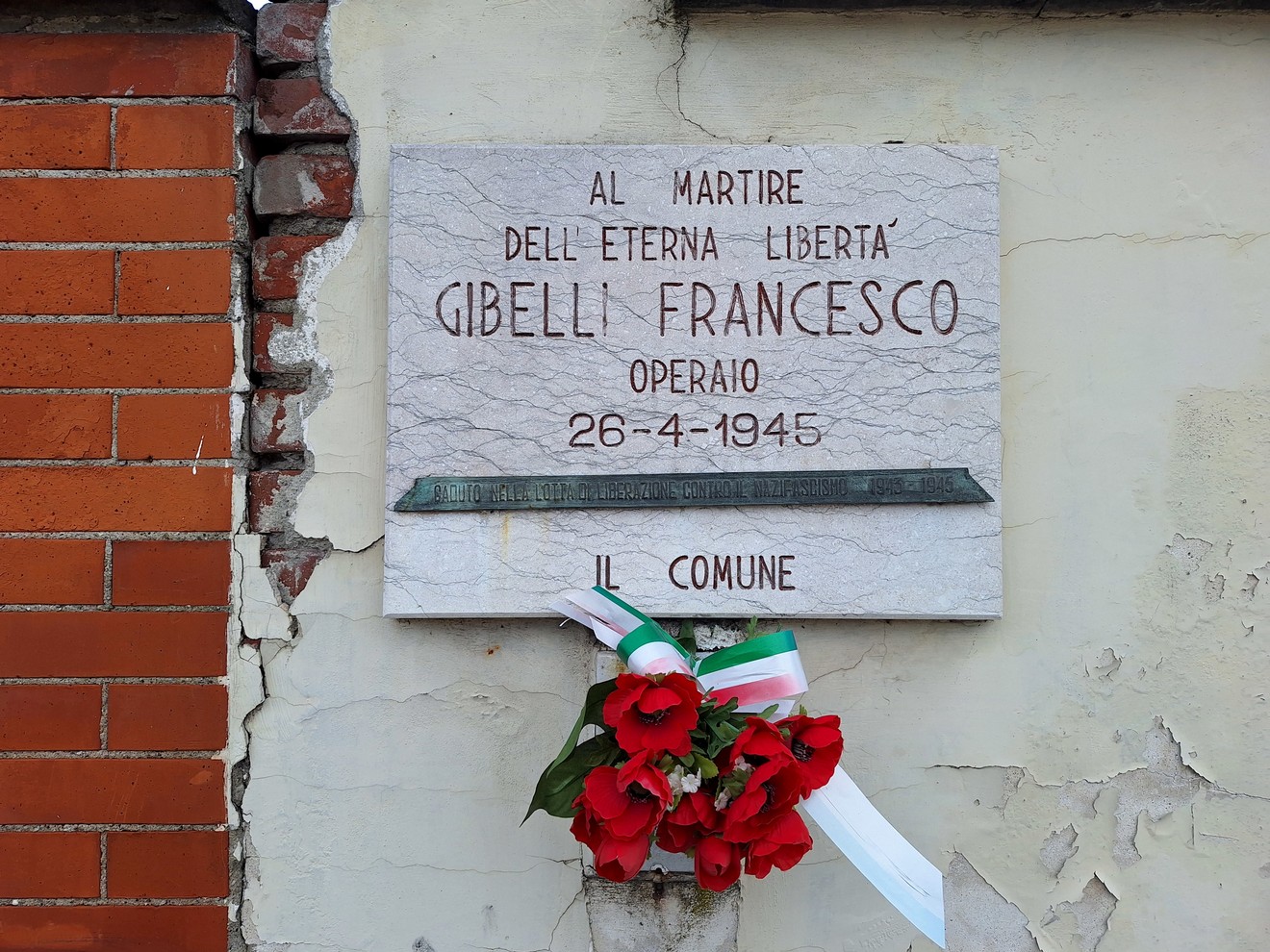 Lapide commemorativa in memoria di Gibelli Francesco, operario, con fiori rossi e nastri tricolore.