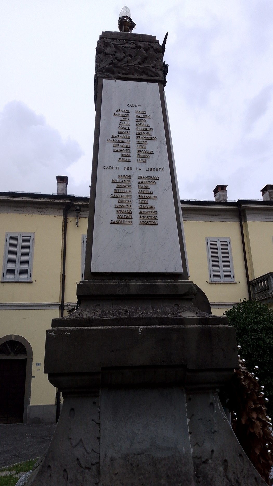 Monumento ai Caduti con iscrizioni commemorative, situato in un contesto urbano, sotto un cielo nuvoloso.