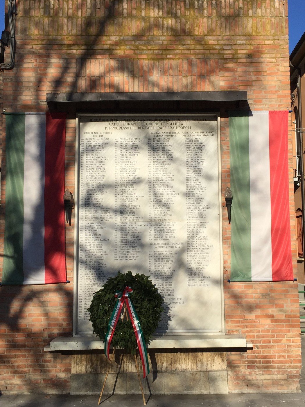 Lapide commemorativa ai caduti di Crespellano, con bandiere italiane e una corona di alloro.