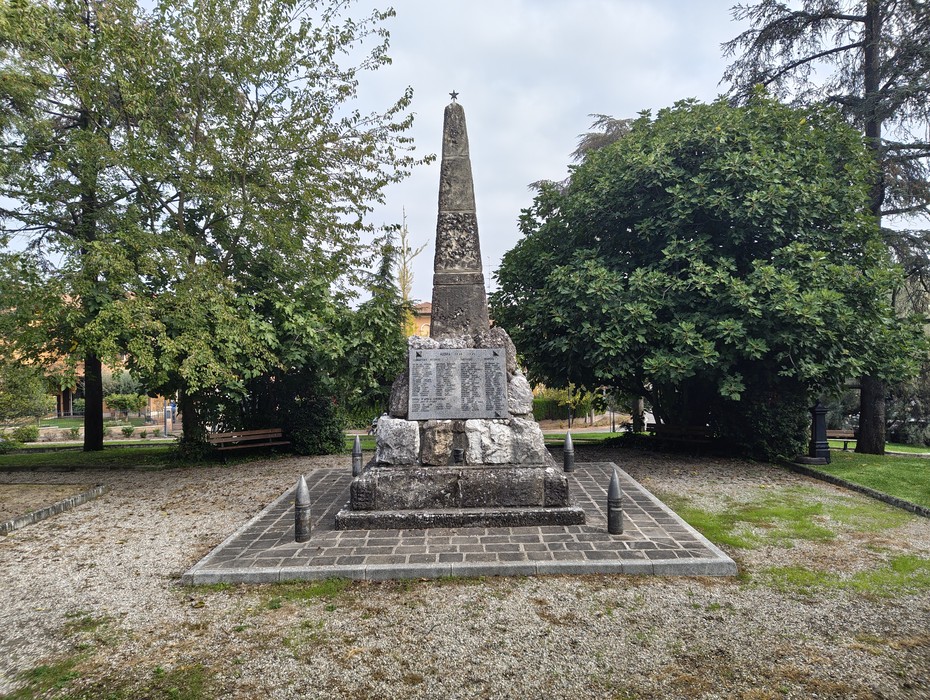 Obelisco in pietra con lapidi incise su base rettangolare, in un'area verde alberata.