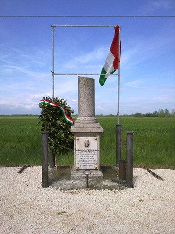 Cippo commemorativo con bandiera italiana e corona d'alloro, immerso nel verde dei campi circostanti.