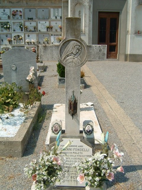 Sepolcro di Don Fondelli, con croce e decorazioni floreali, situato in un cimitero.