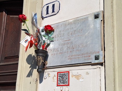 Lapide in marmo su parete esterna, con iscrizione, fiori rossi in un portafiori e codice QR accanto.