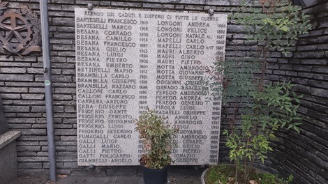 Monumento ai caduti di Cavenago, con nomi incisi su una grande lastra, circondato da piante e muratura in pietra.