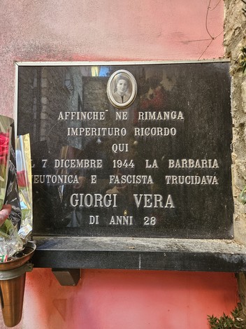 Lapide commemorativa dedicata a Giorgi Vera, con fiori e scritta di ricordo su sfondo roccioso.
