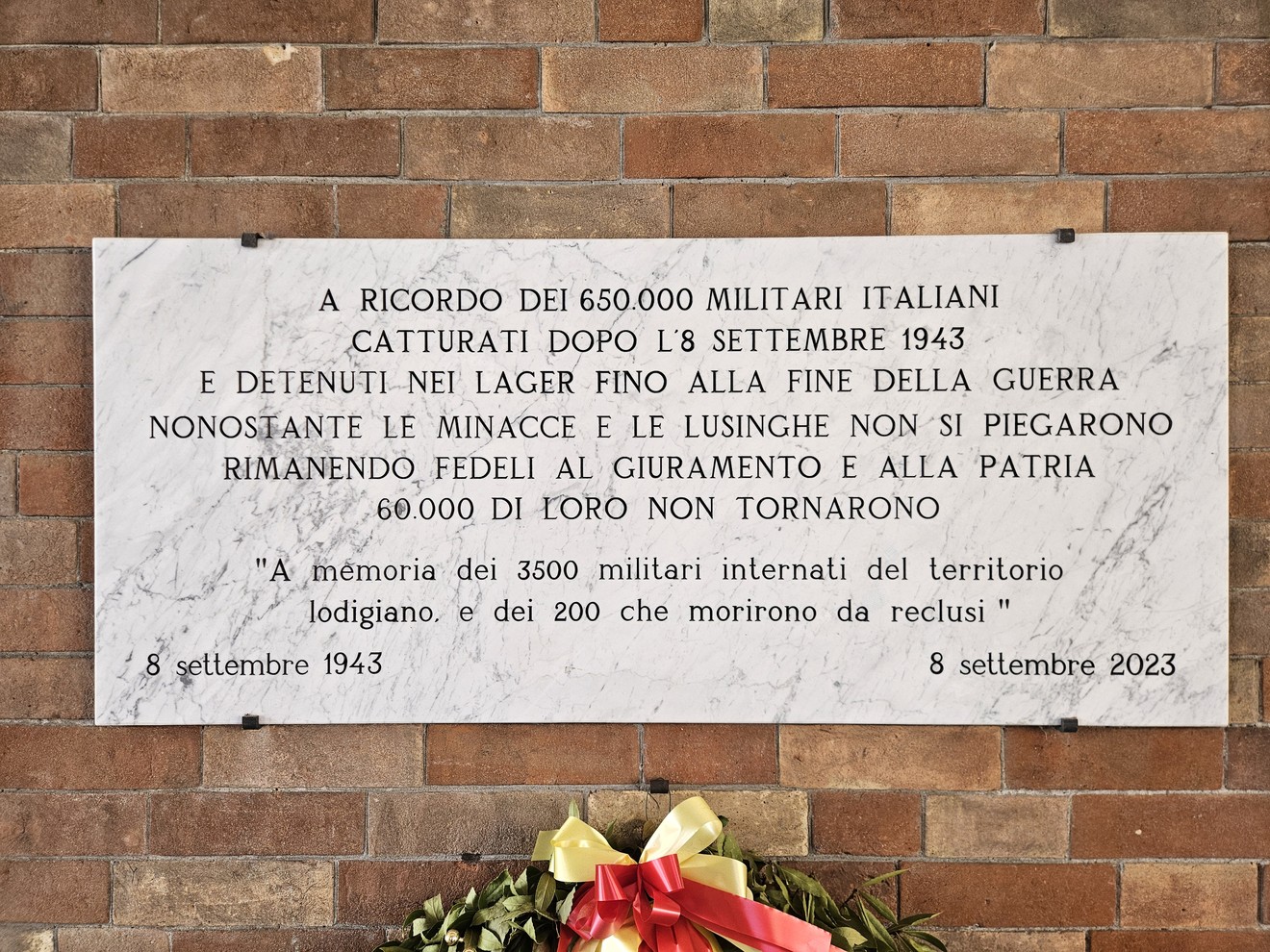 Lapide in marmo su parete di mattoni, con iscrizione e corona di fiori appoggiata sotto.