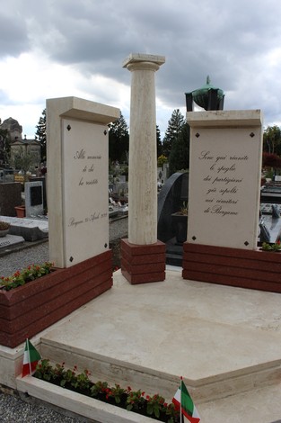 Monumento commemorativo dedicato ai partigiani, con colonne e iscrizioni, circondato da fiori e bandiere.