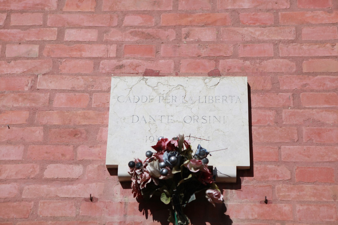 Lapide commemorativa dedicata a Dante Orsini, con decorazioni floreali, su muro di mattoni.