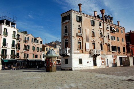 Lapide commemorativa in una piazza storica, circondata da edifici e un cielo azzurro.