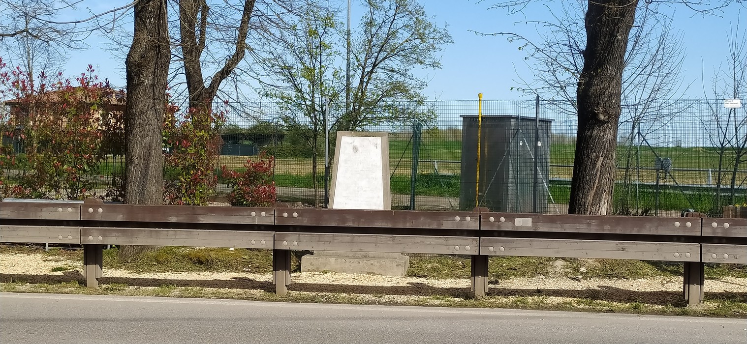 Monumento ai Caduti di Le Budrie circondato da vegetazione e campi verdi in un giorno sereno.
