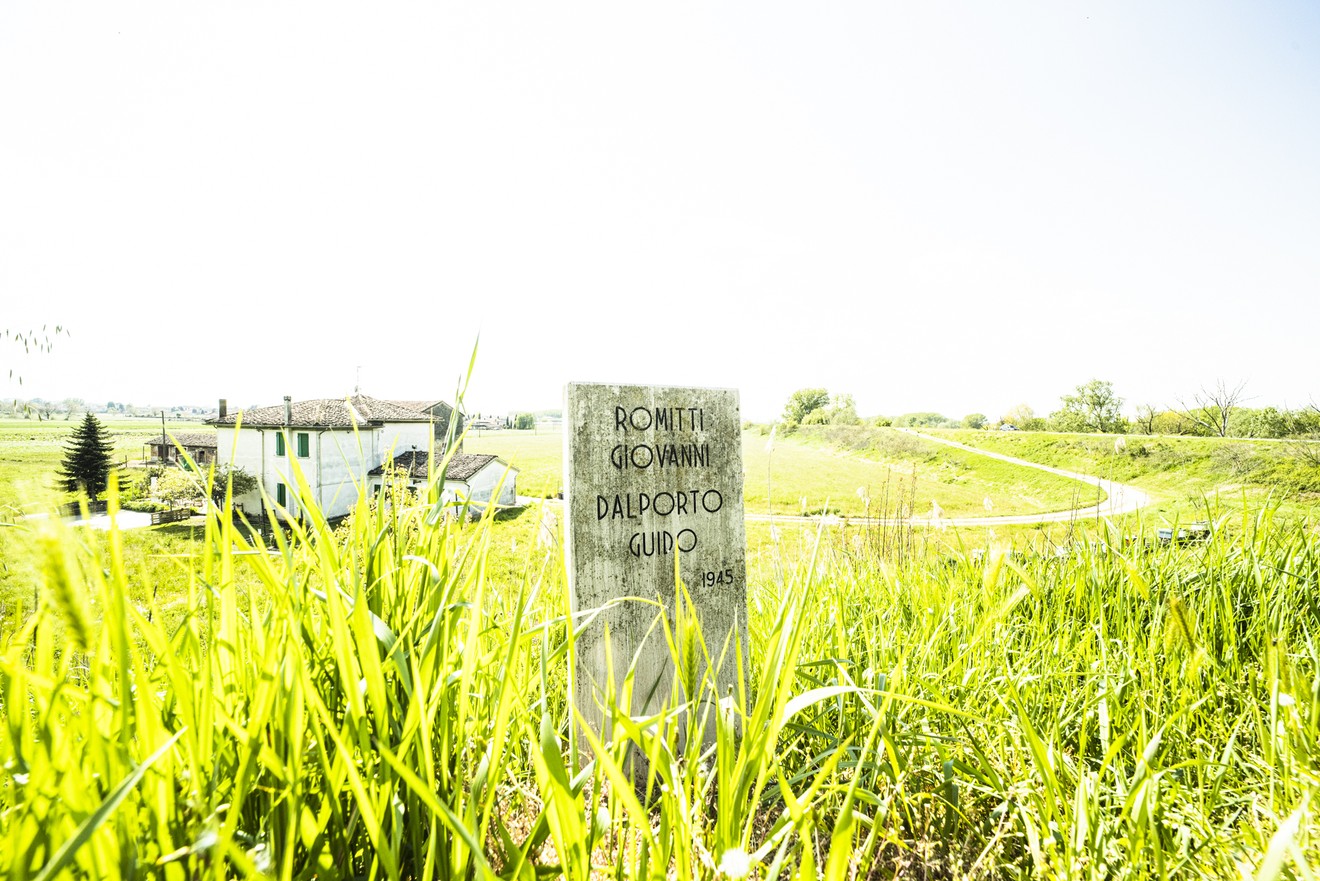 Cippo commemorativo a Romitti e Dalporto, immerso nella vegetazione, con sfondo di campagna.
