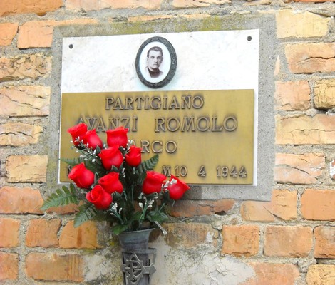 Targa commemorativa di Romolo Avanzi, partigiano, con fiori rossi, posta su un muro di mattoni.