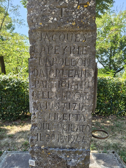 Cippo in pietra con iscrizione dedicatoria, immerso nel verde di alberi e cespugli.