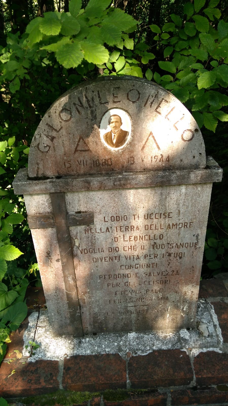 Cippo in pietra con fotografia ovale e iscrizione, circondato da vegetazione.