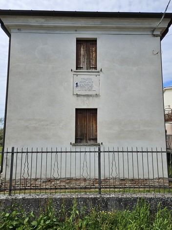 Lapide a Mazzo su un muro di una casa, con finestre chiuse e recinzione in metallo.