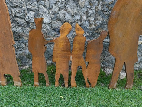Silhouetti di figure umane in metallo patinato, disposte su un prato verde vicino a una parete di pietra.