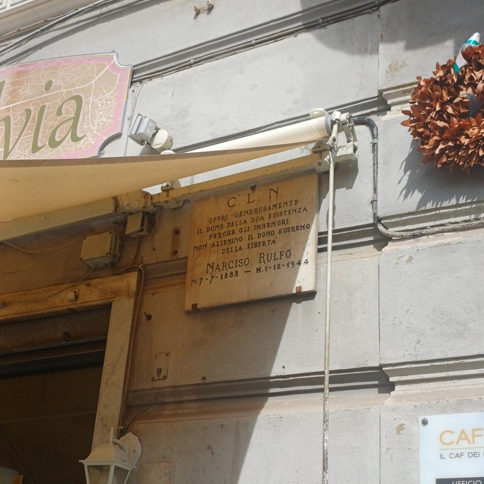 Lapide in marmo con iscrizione, fissata sulla parete esterna di un edificio, sotto una tenda da sole.