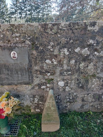 Cippo commemorativo con croce, posizionato accanto a una parete di pietra, circondato da fiori sul prato.