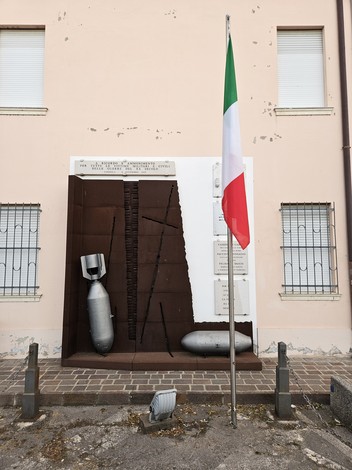 Monumento commemorativo con elementi di artiglieria, dedicato alle vittime militari e civili, e bandiera italiana.