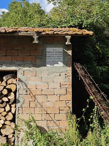 Lapide commemorativa su un muro di mattoni, circondata da vegetazione e legna accatastata.
