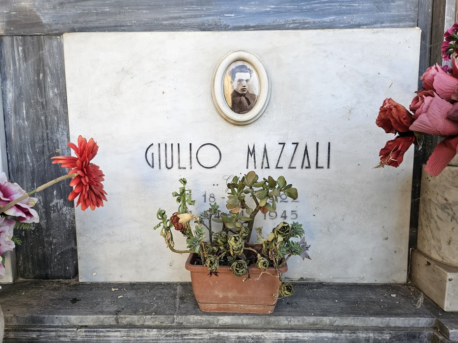Lapide marmorea con foto ovale e nome inciso, decorata con fiori artificiali e una pianta grassa in vaso.