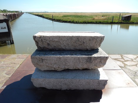 Monumento a Isepetto con blocchi di pietra in primo piano, affacciato su un paesaggio di acqua e vegetazione.