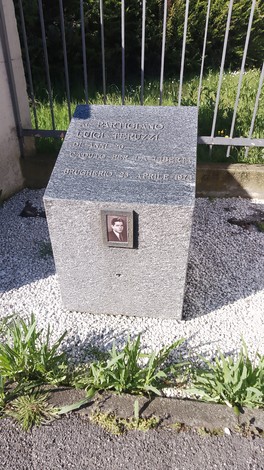 Cippo commemorativo in pietra con un'immagine affissa, circondato da ghiaia e vegetazione.