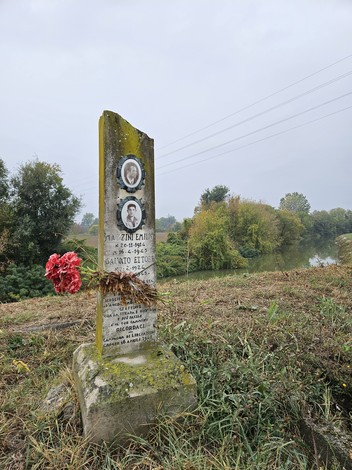 Cippo in pietra con due ritratti fotografici e iscrizione, fiori rossi ai piedi, lungo un canale in campagna.