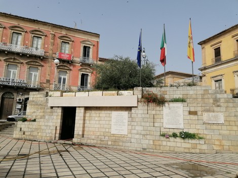 Lapidi commemorative in Piazza del Popolo, con bandiere e una struttura in pietra.