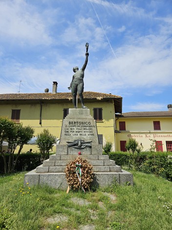 Monumento ai Caduti di Bertonico, con statua eretta su un basamento, circondato da verde e case storiche.